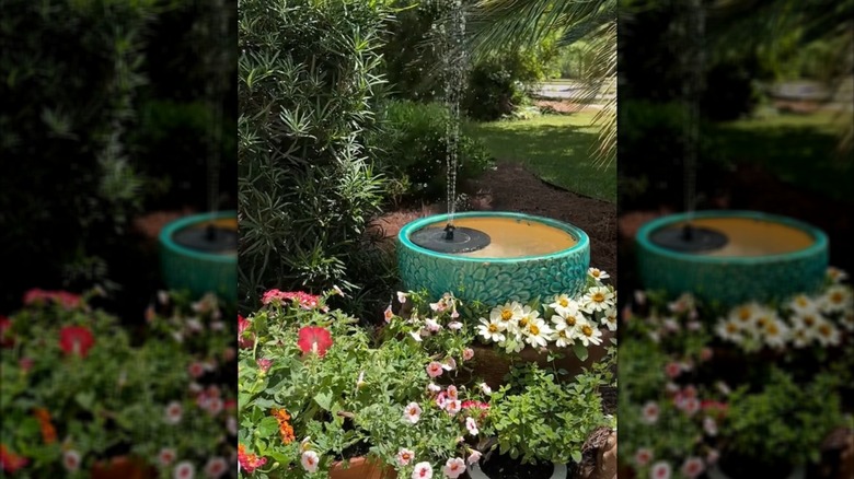 En grønn potte med utvendig blomsterdesign er begravet i en plantekasse, fylt med vann og et solcellebasert fontenetilbehør.