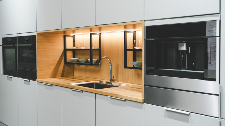 En sølv kjøkkenvask, armatur og hvitevarer satt inn i en benkeplate i tre og bakplate og grå kjøkkenskap.