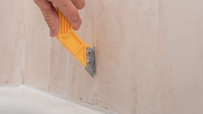 En hånd som bruker et gult caulkverktøy for å fjerne silikon caulk fra mellom fliser.