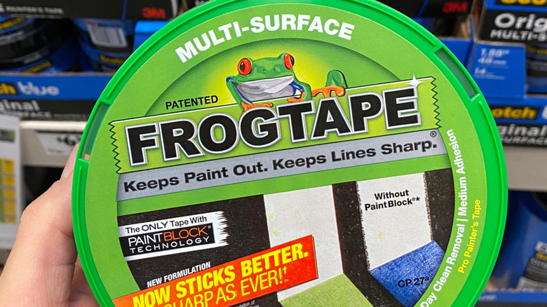 En hånd holder en rull med grønn FrogTape foran en jernvarebutikkhylle.
